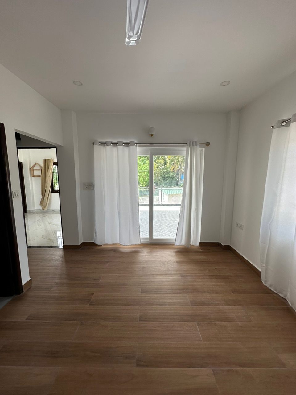 Finished living room with wood-effect floor tiles, white curtains and a sliding door opening to a balcony