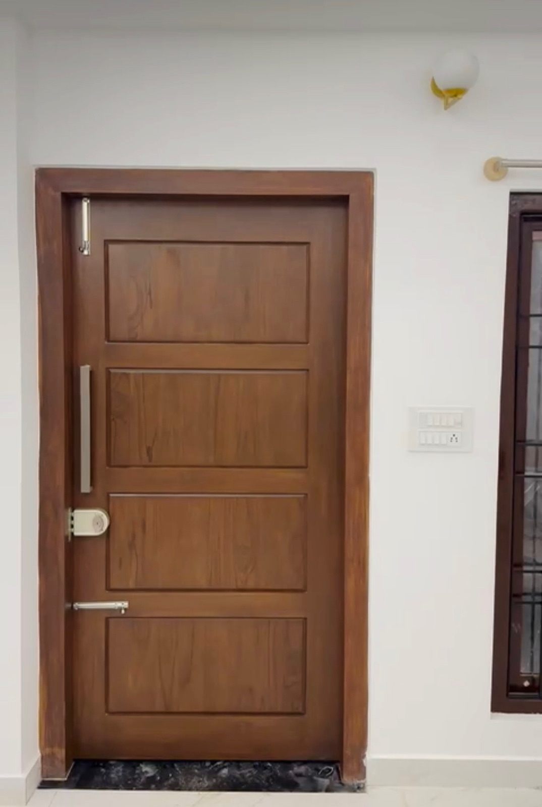 Polished four-panel hardwood main door with a long steel handle and digital lock, set in a matching timber frame