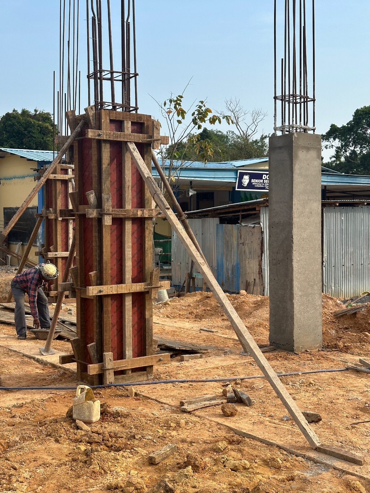 A braced timber column shutter on site next to a freshly stripped concrete column, with a worker in a hard hat